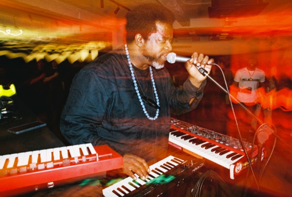 A man in a dark tunic with a string of pale beads sings into a handheld microphone while playing a red keyboard, a second keyboard beside him.
