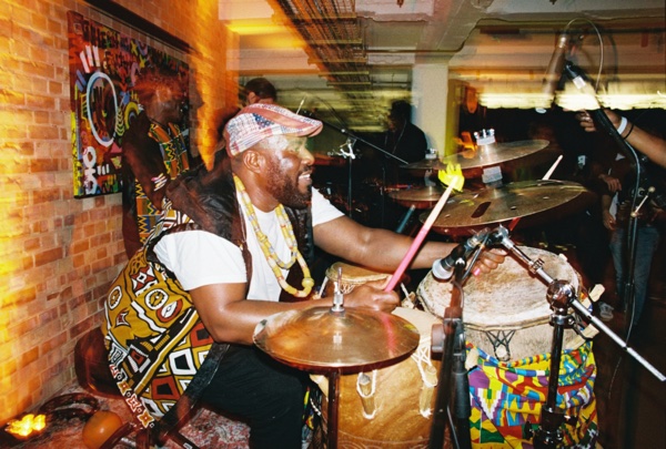 A drummer in a patterned headscarf and a yellow and white wrapper grins mid-strike behind a kit of cymbals and hand drums, in a brick-walled room hung with a painted cloth.