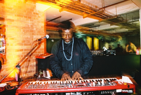 A man in a dark tunic and pale beads plays a red stage keyboard with both hands, head down, in a brick-walled room with a mic stand to his left.
