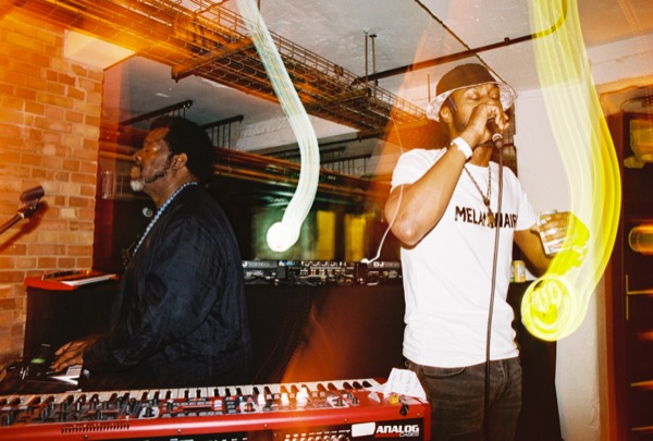 Two performers: one at a red keyboard in a dark tunic, one standing at a microphone in a white printed T-shirt holding a drink. Yellow light trails cross the frame.