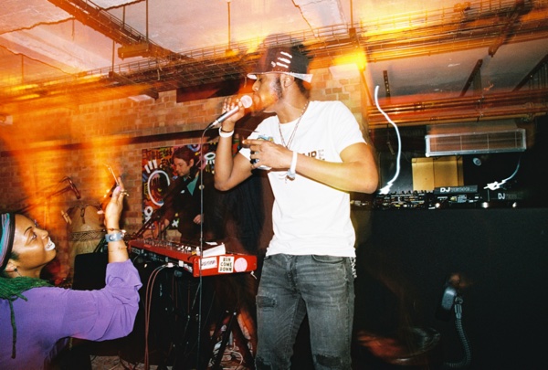 A singer in a white T-shirt and a dark cap sings into a handheld microphone. In the foreground someone in a purple top holds a phone up to film. Orange light trails streak across the frame.