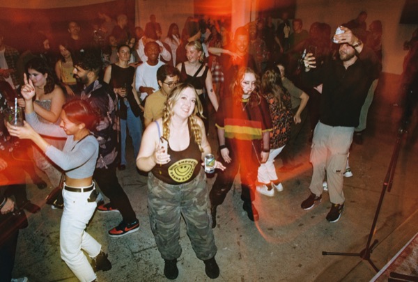 A crowd dances in a concrete-floored room, several people holding drinks and looking towards the camera. A vertical red light streak falls across the middle of the frame.