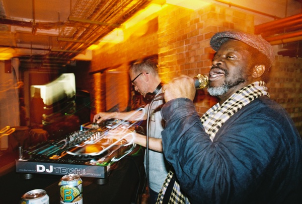 Two men behind a DJ deck: one in glasses and headphones works the mixer while the other, in a flat cap and a checked scarf, laughs into a gold microphone.