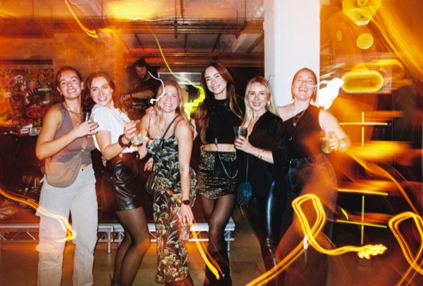 Six people stand together posing for the camera with drinks in hand, in a room with exposed ceiling pipes. Loops of yellow light trail across the frame.