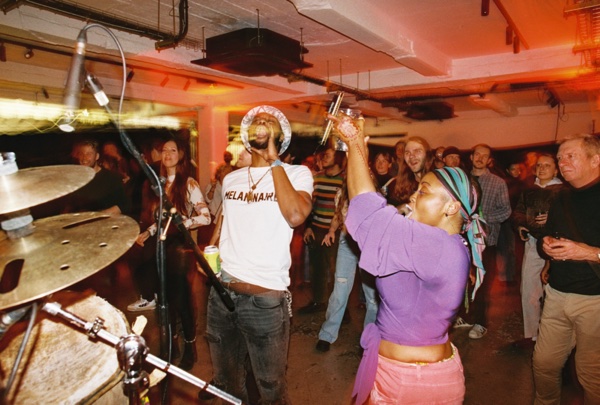 A woman in a purple cardigan raises a drink above her head in a crowded low-ceilinged room. Behind her a man in a slogan T-shirt and a halo-shaped hat dances, with a cymbal in the foreground.