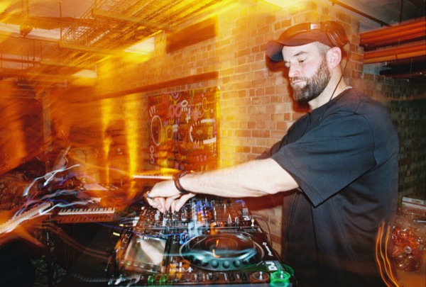 A man in a cap and black T-shirt works a DJ mixer beside a brick wall, arms crossed over the controls. Orange light trails blur the left half of the frame.
