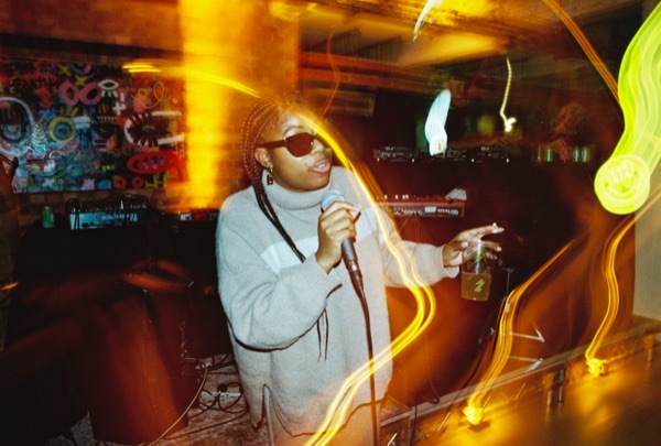 A woman in a pale grey roll-neck jumper and dark glasses, hair in braids, sings into a handheld microphone. Yellow light trails curve across the frame.