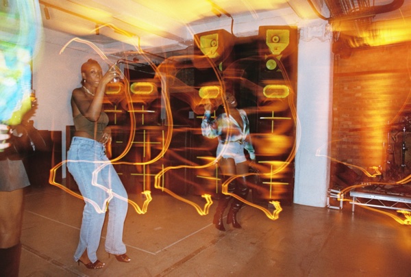 Two women dance in a bare low-ceilinged room with speaker cabinets fixed to the wall, loops of yellow light trailing across the whole frame from a long exposure.