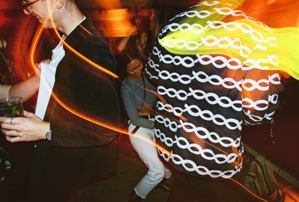 Dancers packed close in orange light: a man in a dark jacket holding a drink at the left, a woman in a grey top behind him, and a large white-patterned coat filling the right of the frame.