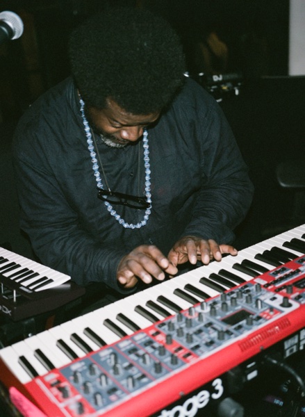 Looking down on a keyboard player with an afro and a long beaded necklace, head bowed and both hands on the keys of a red stage piano stacked above a second keyboard.