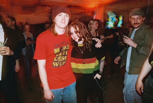 A couple pose arm in arm in a crowded room, one in a brown beanie and a red T-shirt printed with block lettering, the other in a striped red, yellow and green jumper. People stand talking around them in orange light.