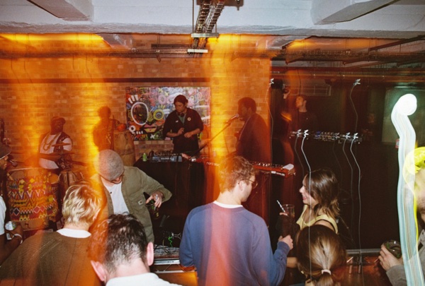 The room seen from the back of the crowd: a band playing on a low stage under a painted canvas on a brick wall, a drummer at the left, and people standing with drinks in the foreground. Orange light streaks down the left side.