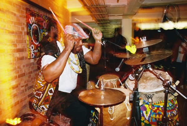 A drummer in a white vest and a brightly patterned wrap and cap raises both sticks over a kit of cymbals and hand drums, a painted canvas hanging on the brick wall behind him.