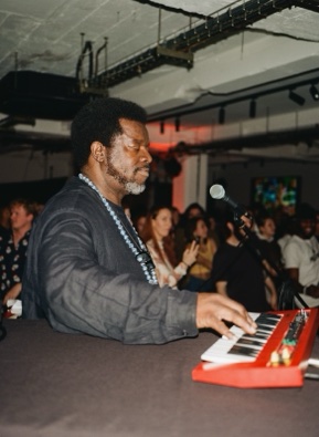 A keyboard player in a dark grey tunic and long strings of blue beads plays a red synthesiser with his eyes down, a crowd and a lit ceiling grid behind him.