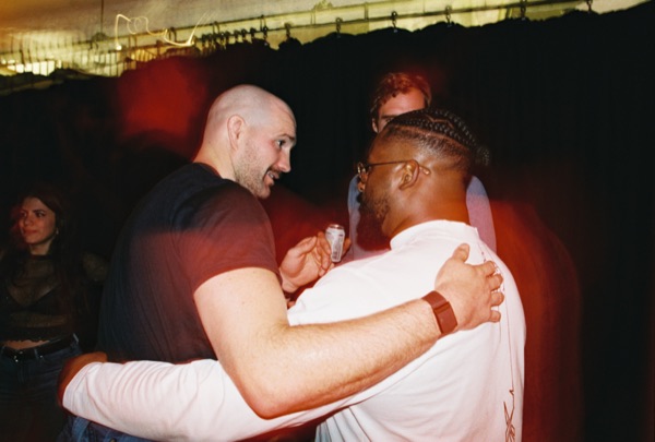 Two men embrace and talk closely at a party, one in a dark T-shirt, one in a white top with braided hair. Others stand behind against a dark curtain.