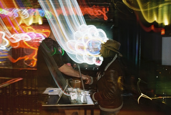 Two people lean over a table in a dark bar, one of them in a wide-brimmed hat, while looping trails of coloured neon light swirl across the whole frame.