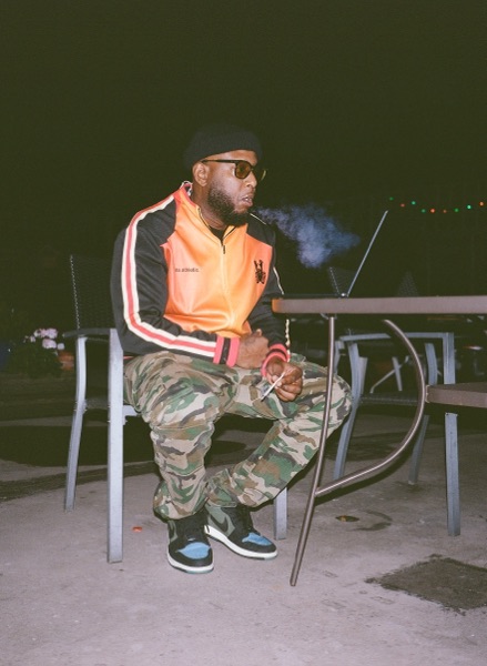 A man in an orange and black track jacket, beret and sunglasses sits on a metal chair outdoors at night, exhaling smoke, a table and string lights behind him.