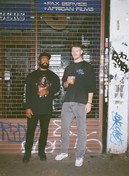Two men stand for a portrait in front of a graffitied shop shutter at night, one in a black printed T-shirt, one in a dark sweatshirt holding a drink. A sign above the shutter advertises services in blue and white lettering.