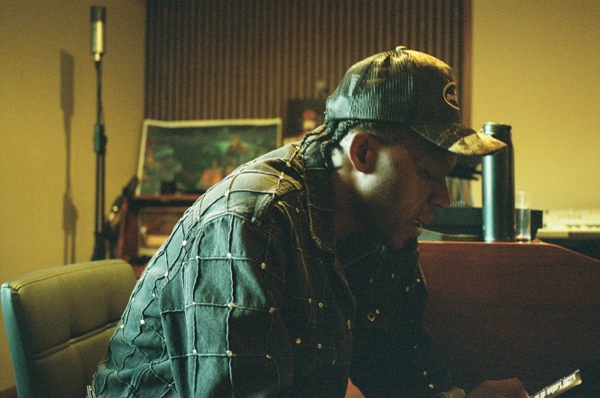 A man in a camouflage cap and a studded denim jacket leans forward in a warm-lit room with his head down, a framed painting and a microphone stand behind him.