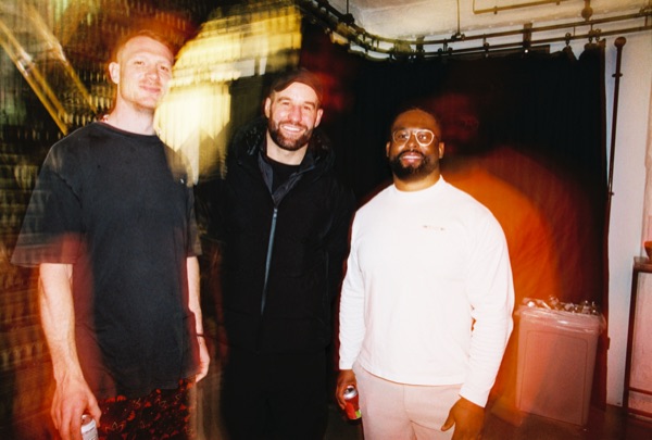 Three men stand shoulder to shoulder for a portrait, smiling at the camera. A warm blur and light leak softens the edges of the frame.