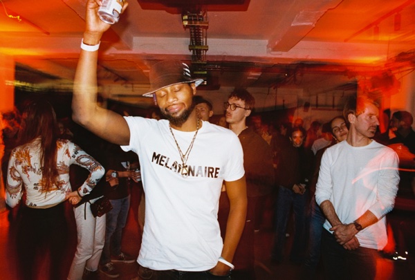 A man in a slogan T-shirt, a gold chain and a pale cap raises a can above his head in a crowded room, with orange light leaking across the frame.
