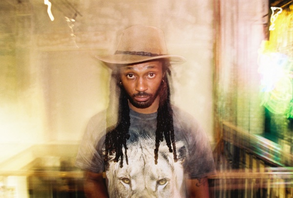 A portrait of a man in a wide-brimmed felt hat with long locs, wearing a T-shirt printed with a lion's face, lit by a warm haze with green light behind him.