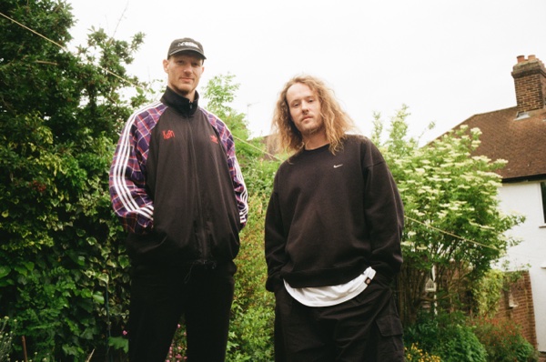 Two men stand side by side in a garden, photographed from low down against a pale overcast sky, one in a dark cap and a track jacket with tartan sleeves, the other with long fair curls in a plain black sweatshirt. An overgrown hedge, a flowering shrub and the corner of a house are behind them.