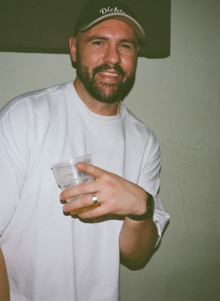A man in a dark cap and a loose white T-shirt smiles at the camera, holding a clear plastic cup of drink towards the lens, against a plain pale green wall.