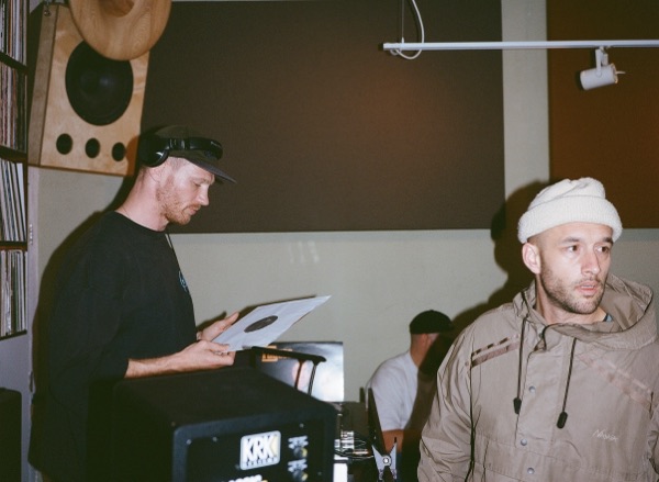 A man in headphones and a cap holds a record sleeve over a speaker cabinet while another man in a white beanie and pale jacket looks off to the right.