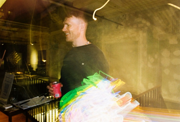 A man in a black T-shirt laughs, looking off to one side, a red drinks can in his hand as he leans on a railing. Green and yellow light trails blur across the lower half of the frame.