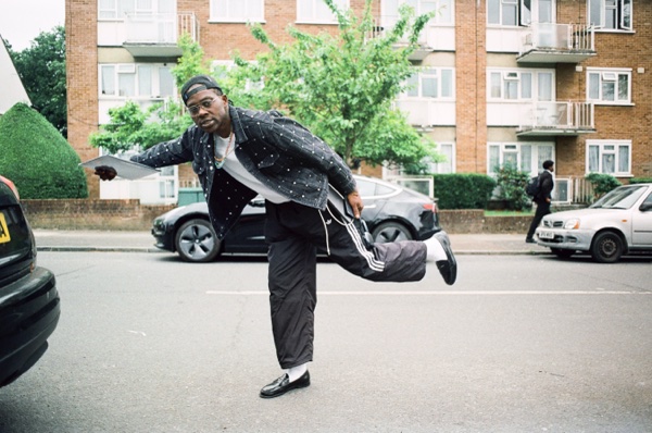 A man in a spotted black jacket, dark glasses and a headscarf leans into a dance step in the middle of a suburban street, one leg kicked out behind him, with parked cars and a brick block of flats behind.