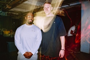 Two men stand for a portrait indoors, one in a pale sweatshirt and glasses, one taller in a dark T-shirt. A soft light leak washes vertically through the middle of the frame.