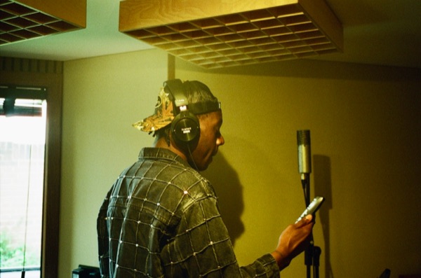 A man in a checked shirt and headphones stands facing a large studio microphone on a stand, holding a phone in one hand, under a slatted wooden ceiling panel.