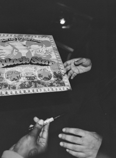 Black and white frame of two pairs of hands over a record sleeve. One hand holds a marker pen; the sleeve carries large display lettering and has been signed across the front.