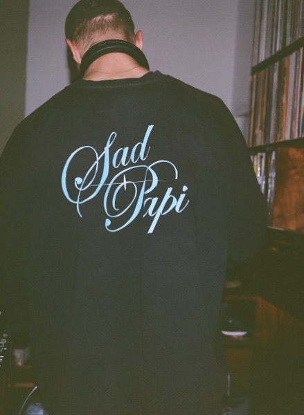 A man photographed from behind wearing a black long-sleeved top with flowing script lettering printed across the back, standing in front of shelved records.