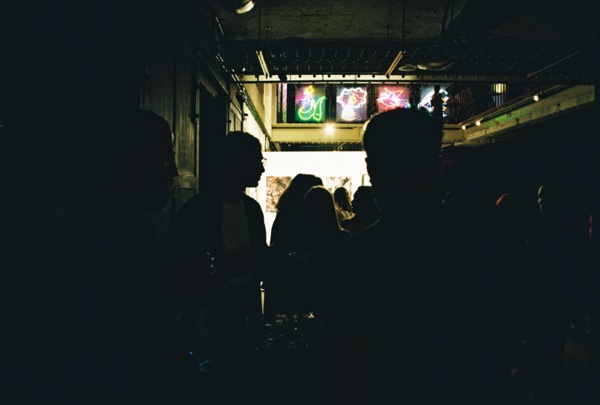 A dark room of silhouetted figures. At the far end a lit projection of four bright panels is the only source of light.