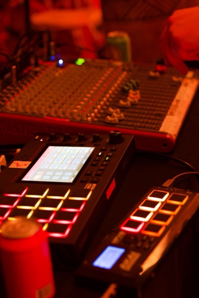 Close view of a drum sampler and a mixing desk under red light, the sampler's pads glowing red and orange, a small screen showing a grid of cells.