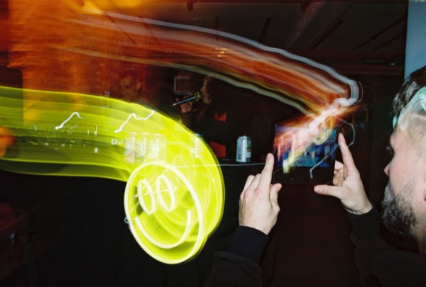 A man holds a phone up to film. A thick yellow light trail loops across the frame in front of him and coils into a spiral.