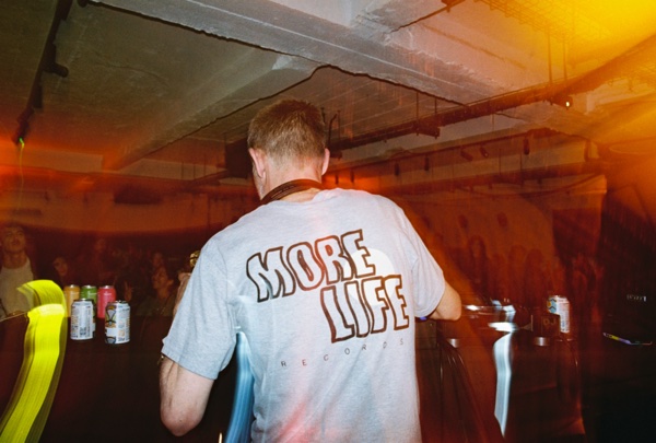 Seen from behind, a person in a grey slogan T-shirt stands at the decks. Cans sit on a ledge to the left and a warm orange light leak washes the right of the frame.