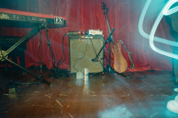 Stage equipment at rest in front of a red curtain: a keyboard on a stand, a guitar amplifier, an acoustic guitar, and cables run across a wooden floor. A pale light trail curves in from the right.