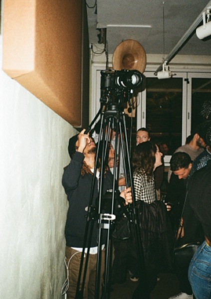 Two people adjust a large cine camera mounted on a tripod beside a draped wall, with a small crowd standing and talking behind them.
