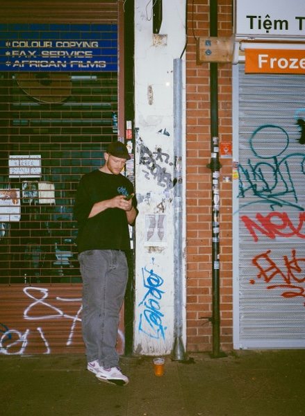 A person in a black T-shirt, grey cargo trousers and white trainers stands looking at their phone against a shuttered shopfront covered in graffiti, beside a brick pillar.