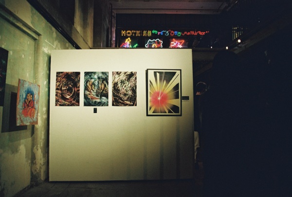 Prints hung on a white display board in a dark room: three swirling abstract photographs in a row and a larger framed print of a red and yellow starburst. Neon lettering glows on the wall above and a small painting hangs on the bare wall at the left.