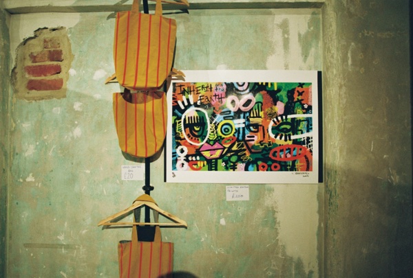 A print of a densely painted canvas of faces, eyes and lettering, reading INHERIT THE EARTH across the top left, hangs on a peeling plaster wall with a handwritten price card beside it. Orange striped tote bags hang from a rail to the left.
