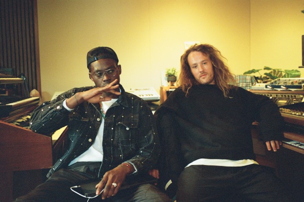 Two men sit side by side in a studio, one in a black cap and a studded denim jacket making a two-finger sign, the other with long fair hair in a black sweatshirt. A keyboard, a plant and shelves of equipment fill the wall behind them.