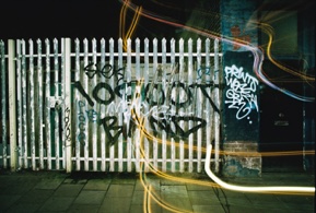 A white picket fence covered in black spray-painted tags at night, with light trails streaking across the pavement in front of it.