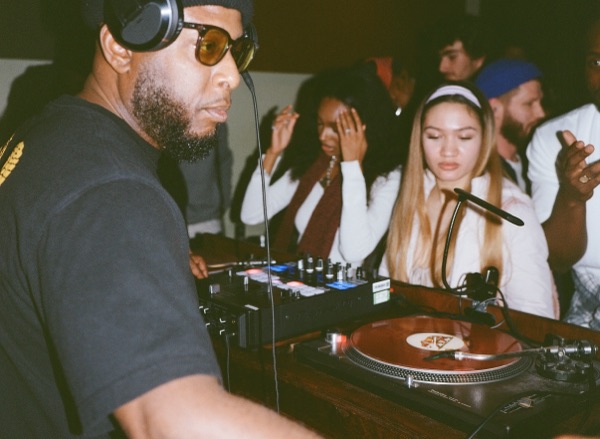 A DJ in headphones and yellow-tinted glasses works the turntables in the foreground. A row of people stand watching from the far side of the booth.