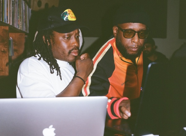 Two men lean in towards an open laptop, lit from below by the screen. One wears a cap and white T-shirt, the other an orange track jacket and dark glasses.