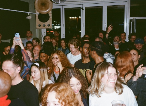 A dense crowd fills a bright room with folding glass doors along one wall. One person holds a phone overhead to film. A large horn speaker is mounted on the wall above.