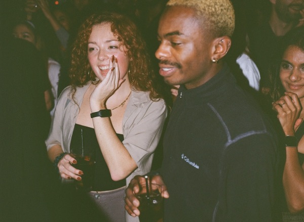 A woman with curly red hair laughs with one hand raised to her face, beside a man with close-cropped bleached hair in a black fleece. A packed room behind them.